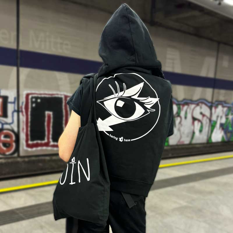 Ein junger Mann steht mit dem Rücken zum Betrachter auf einem U-Bahnsteig. Er trägt einen schwarzen Kapuzenpullover mit einem Auge als Aufdruck.